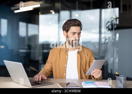 Businessman sitting at a sleek desk, using a laptop and holding a digital tablet, focused on a project in a modern office featuring bright glass walls and contemporary design Stock Photo