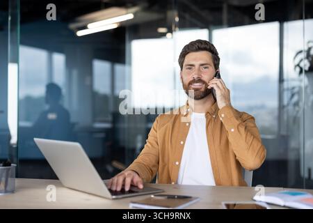 Smiling businessman multitasking at his modern office desk, talking on a mobile phone while working on a laptop, showcasing efficiency and productivity in a bright, professional environment Stock Photo