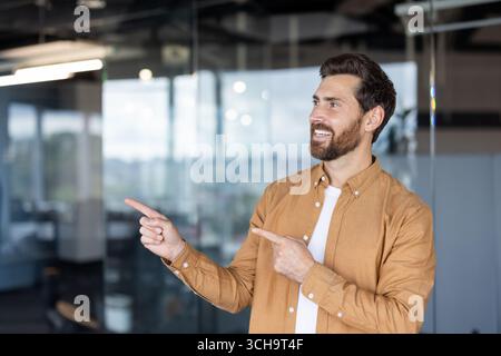 Businessman is smiling and pointing with both index fingers towards the side in a modern office environment, suggesting a new opportunity or highlighting important information Stock Photo