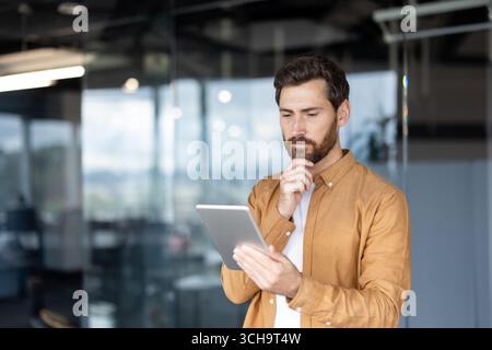 Focused entrepreneur using tablet, touching his chin with hand, analyzing data, working on new project in modern office space, developing innovative business strategy Stock Photo