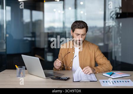 Focused businessman holding eyeglasses and reviewing financial documents, working with laptop, calculator, and charts at his desk in a bright, modern office Stock Photo