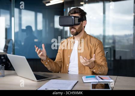 Office worker using vr headset is interacting with virtual reality in modern office, gesturing with his hands while sitting at desk with laptop and documents Stock Photo