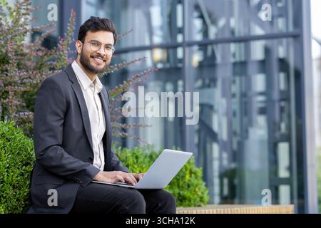 Young businessman is sitting outside with his laptop, working remotely near office building, enjoying fresh air and sunshine while staying connected and productive Stock Photo