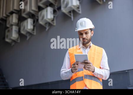 Building maintenance technician wearing safety helmet and vest, using digital tablet and inspecting air conditioning units on building exterior Stock Photo