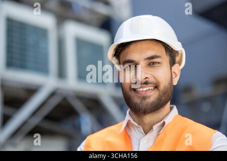 Indian engineer wearing a hardhat and safety vest smiles confidently in an industrial setting, possibly a warehouse or factory, with hvac units in the background Stock Photo