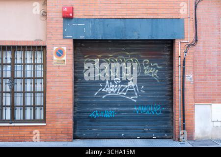 Puerta de garaje azul verdosa con grafiti  firmas, fachada de ladrillo visto con ventana, cableado eléctrico expuestp, alarma de incendios roja arriba Stock Photo