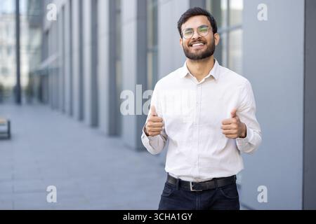 Happy businessman showing thumbs up gesture while standing near modern office building, expressing positivity and success in business career Stock Photo