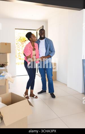 Happy African American couple embracing in new home surrounded by moving boxes, celebrating together. Homeownership, relocation, unpacking, love, rela Stock Photo