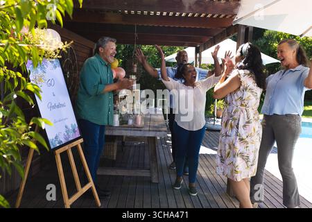 Diverse senior friends celebrating birthday outdoors, laughing and raising hands in joy. Celebration, happiness, party, elderly, gathering, festive Stock Photo