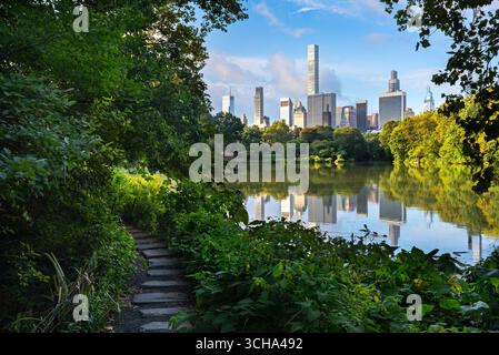 Central Park summer morning with stone path and lake reflecting Midtown Manhattan skyline. New York City Stock Photo