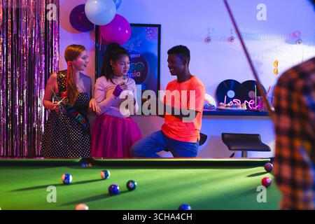 Teenage diverse friends celebrating birthday party playing pool and chatting in vibrant room, at home. Celebration, fun, friendship, leisure, entertai Stock Photo