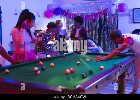 Diverse teens enjoying birthday party playing pool and socializing in festive room, at home. Celebration, diverse teenagers, billiards, diverse friend Stock Photo