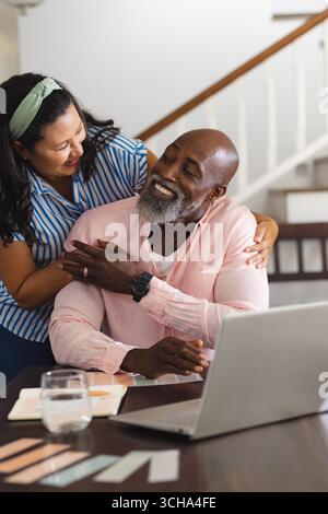 African American senior diverse couple smiling and embracing while using laptop at home. Lifestyle, technology, communication, diverse senior friends, Stock Photo