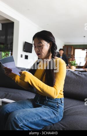 Asian teen reading letter on couch, expressing surprise and excitement at home. Teenager, emotion, communication, revelation, living room, leisure Stock Photo