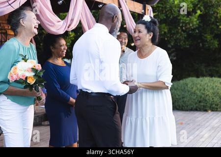 At wedding, celebrating in garden, senior diverse couple holding hands with friends, smiling. celebration, friendship, elderly, happiness, community, Stock Photo