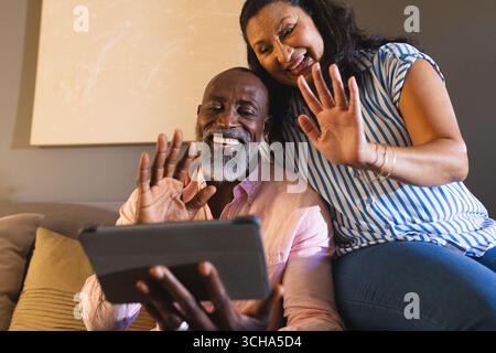 Smiling senior diverse couple video chatting on tablet, waving at home. Elderly, communication, technology, connection, digital, lifestyle Stock Photo