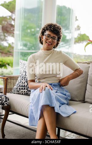 Smiling woman sitting on sofa in bright, modern living room, enjoying moment, at home. Relaxation, leisure, happiness, lifestyle, comfort, interior Stock Photo
