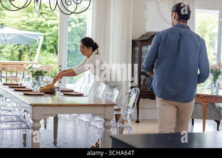 Woman setting elegant dining table with flowers while man observing in bright room, at home. Table setting, elegance, lifestyle, home decor, interior Stock Photo