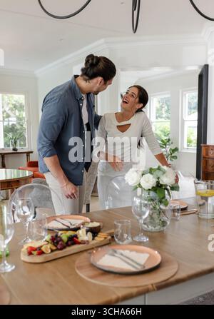 Couple laughing together at dining table with elegant table setting and flowers, at home. Romance, celebration, happiness, laughter, intimacy, tablewa Stock Photo