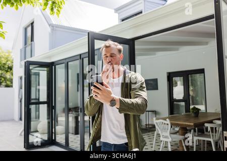 Man outdoors using smartphone, surprised and smiling in modern home patio. Technology, communication, lifestyle, happiness, leisure, relaxation Stock Photo