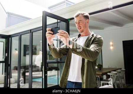 Young man using smartphone to capture moments at home, smiling and relaxed. Technology, casual, lifestyle, communication, relaxation, cheerful Stock Photo