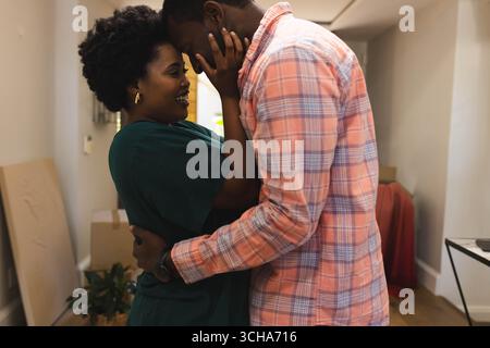 Hugging african american couple standing in hallway at home, with moving boxes and potted plant. romance, intimacy, interior, warmth, connection, deco Stock Photo