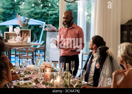 At wedding reception, man giving heartfelt speech, guests listening attentively around table. Celebration, toasting, marriage, happiness, event, socia Stock Photo