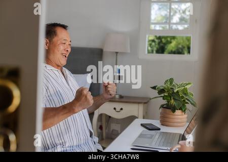 Senior man celebrating success while using laptop at home office desk. Elderly, technology, achievement, working, happiness, win Stock Photo