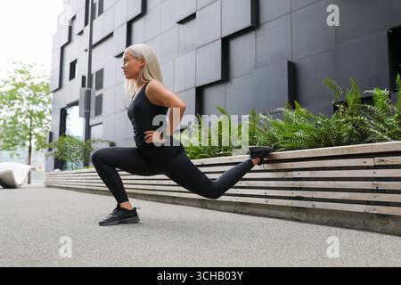 Active woman exercising outdoors with urban background, performing leg stretch lunge Stock Photo