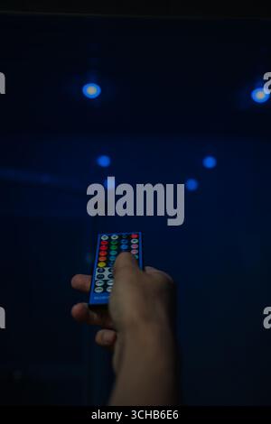 Close-up hand holding RGB remote control, poised to adjust smart lighting system, changing ambiance of room or setting specific mood with colorful led Stock Photo