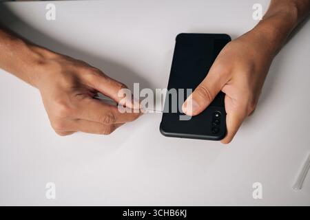 Close-up of male hands utilizing sim ejector tool for removing sim card tray from a smartphone, conducting mobile phone repair on white table. Stock Photo