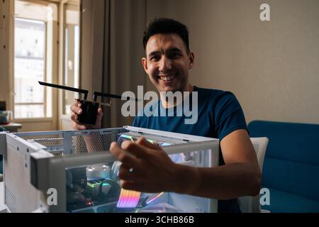 Friendly computer technician male smiling looking camera during installing wi-fi module into custom-built desktop computer with RGB lighting. Stock Photo