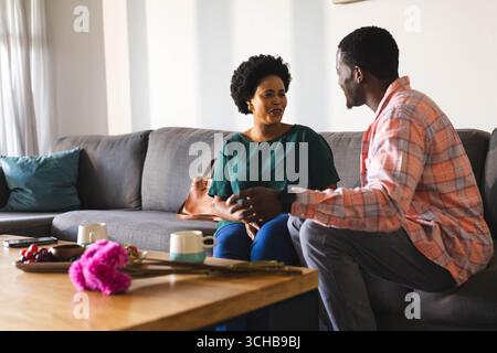Talking African American friends sitting on sofa in living room, with mugs and cherries on table. Collaboration, companionship, cozy, homey, modern, w Stock Photo