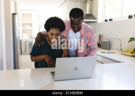 Laughing African American couple reacting at kitchen island, with open laptop and fruit bowl. Contemporary, intimacy, technology, domestic, cheerful, Stock Photo