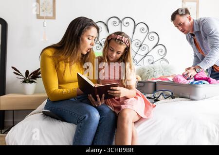Mother and daughter reading book on bed while father packs suitcase nearby. Family, parenting, bonding, leisure, travel, packing Stock Photo