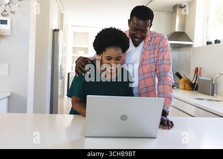 Leaning African American couple laughing while using laptop on kitchen island, with hanging pots. Contemporary, lifestyle, minimalist, domestic, colla Stock Photo