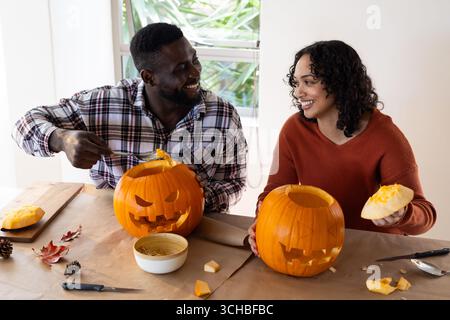 Halloween time, smiling multiracial couple carving pumpkins at home. festival, togetherness, seasonal, decoration Stock Photo