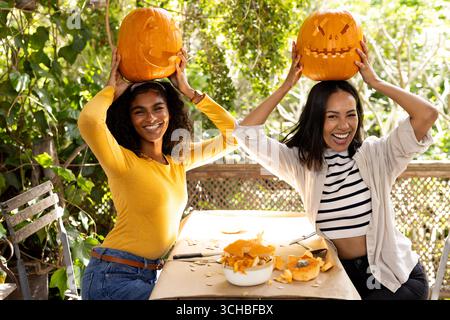 Halloween time, smiling multiracial female friends holding carved pumpkins on heads on deck. halloween, on deck, festive, friendship Stock Photo