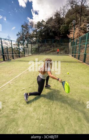 A young interracial couple plays padel on an outdoor court in the countryside of a Catalan village, with her returning a padel ball with a forehand. Stock Photo