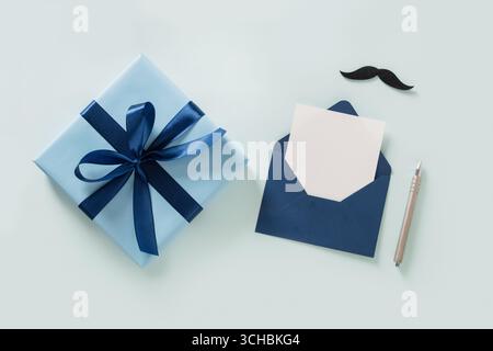 Blue gift box, moustache, white blank in envelope, and pen on pastel blue background. Invitation. Happy Fathers Day. November concept. View from above Stock Photo