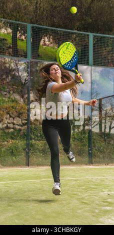 A young and strong athlete playing padel, jumping to hit the ball with a racket and looking very focused and energetic on an outdoor green court. Stock Photo