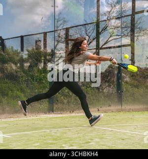 Young sporty woman in mid-jump to return and hit a paddle ball on an outdoor green court in the mountains. Stock Photo