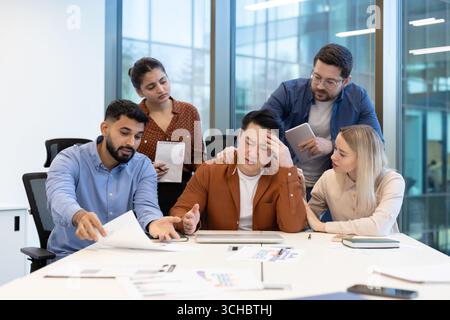 Diverse business team showing support to a stressed asian colleague during a challenging office meeting, demonstrating empathy and teamwork in a diffi Stock Photo