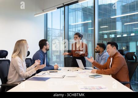 Young businesswoman is leading a meeting in a modern office, presenting to her diverse team who are clapping and celebrating their success Stock Photo