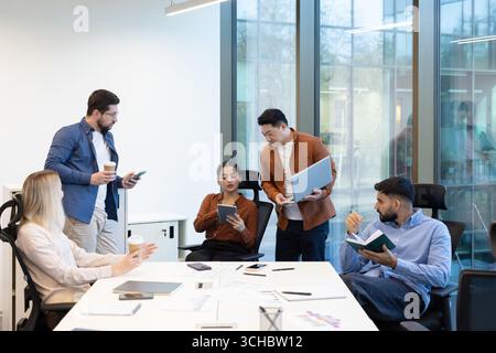 Business team collaborating on a project, using laptop, tablet, and notebook, having a discussion in a modern office, working together to achieve comm Stock Photo