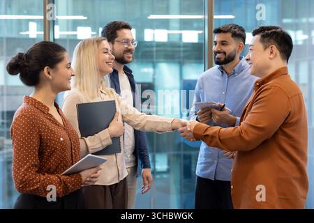 Two business professionals are shaking hands, finalizing a successful deal or partnership, surrounded by their diverse team in a modern office environ Stock Photo