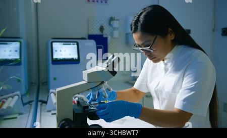A woman researcher in a lab coat analyzes samples using a microscope while wearing gloves in a modern laboratory. Stock Photo
