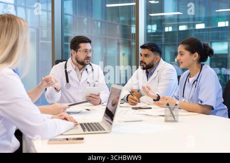 Doctors and nurses sitting around a table in a modern hospital conference room, discussing patient cases and collaborating on treatment plans using a Stock Photo