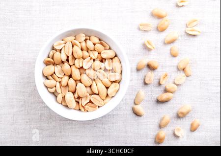 Roasted peanuts in a white bowl on linen fabric. Ready-to-eat crunchy snack food made from fruits of Arachis hypogaea. Light golden snack nuts. Stock Photo