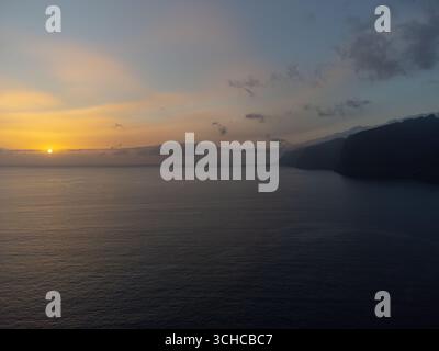 Sun setting over the Atlantic Ocean near Tenerife, Spain, casting warm hues across the sky. The sun is partially obscured by distant clouds Stock Photo